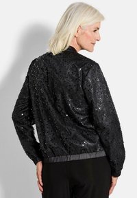 Veste bomber noire avec une surface ornée de sequins, des poignets et un ourlet élastiques, et une texture satinée lisse. Conçue pour un ajustement décontracté.