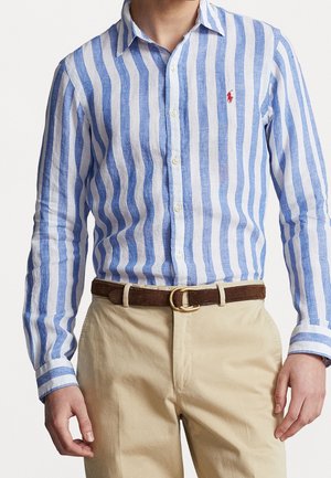 Homme portant une chemise à boutons rayée verticalement bleue et blanche, un pantalon beige et une ceinture marron avec une boucle dorée.