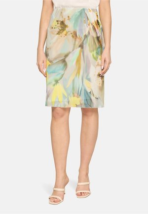 Vrouw draagt een pastelkleurige bloemige kokerrok tot op de knie met beige open-toe sandalen met hak, staand tegen een witte achtergrond.