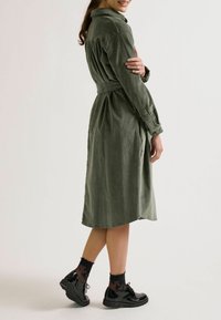 Eine Frau in einem olivgrünen Kleid mit einem taillierten Bund steht seitlich, lächelt und trägt schwarze Schuhe sowie gemusterte Socken.