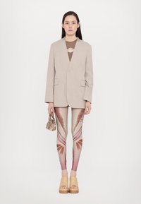 Beige blazer over brown top, patterned leggings with pink gradients, wooden clog sandals, holding a patterned mini bag. Simple background.