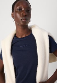 T-shirt in cotone blu navy con logo "TOMMY JEANS", abbinata a un gilet bianco e soffice drappeggiato sulle spalle. Tessuto morbido e design aderente.