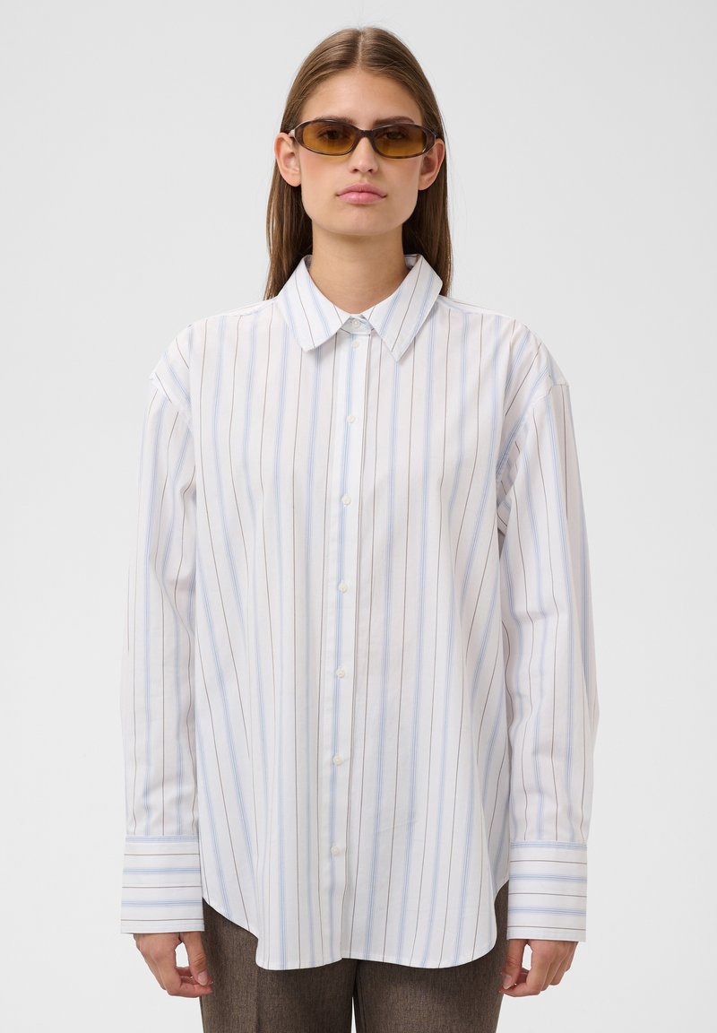 Jeune personne portant des lunettes de soleil ambre et une chemise blanche à manches longues avec des rayures verticales bleu clair et marron, debout devant un fond uni.