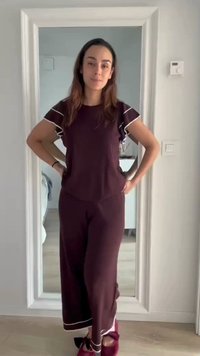 Femme en tenue d’intérieur violet foncé avec bord blanc, debout, mains sur les hanches, devant un miroir en pied dans une pièce minimaliste.