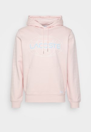 Jasnoróżowa bluza z kapturem z przednią kieszenią, niebieskim logo Lacoste i grafiką mapy świata na piersi oraz wzorem w kratkę na rękawach.