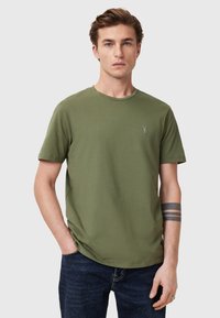 Olivi vihreä lyhythihainen t-paita pehmeästä puuvillasta, jossa on pyöreä kaula-aukko ja pieni brodeerattu logo rinnassa, yhdistettyinä tummiin farkkuihin.