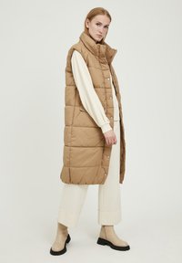 Long gilet matelassé de couleur beige chaud avec un col haut, des boutons pression et des poches latérales, associé à un haut crème à manches longues et des bottes.