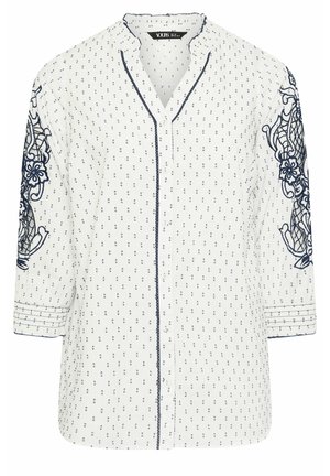 White blouse with navy blue polka dots, featuring embroidered floral design on sleeves, notch collar, and button front. ¾ sleeves with decorative stitching.