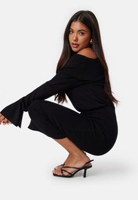Abito nero a coste con design off-the-shoulder e maniche a campana. Silhouette aderente di lunghezza midi, abbinato a sandali neri con tacco sottile.