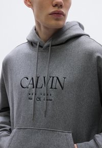 Szürke kapucnis pulóver fekete hímzéssel, amelyen a "CALVIN NEW YORK 1968 CK" felirat olvasható, elöl zsebfedeles és húzózsinóros kapucnival.