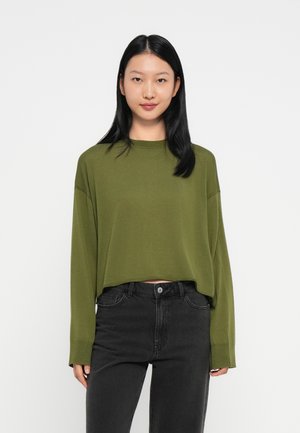 Jeune femme avec des cheveux noirs raides portant un pull court à manches longues de couleur vert olive et un jean noir taille haute sur fond blanc.