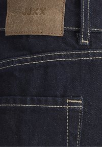 Donkerblauwe denimstof met contrasterende beige stiksels, een riemlus en een bruine leren patch met de opdruk "JJXX" op de tailleband.