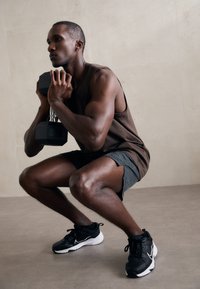 Mançá de halter preto segurada perto do peito por uma pessoa de um top de treino castanho e calções cinzentos, agachando-se num chão cinzento claro. Sapatos de treino pretos usados.