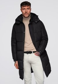 Sort polstret frakke med hætte, der når til midten af låret, med lynlåslukning, knapdetaljer og elastiske manchetter, båret over en beige turtle-neck.