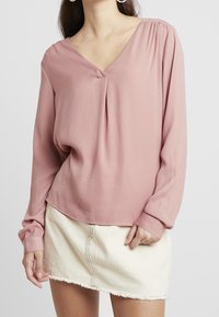 Blusa rosa a maniche lunghe con scollatura a V, spalle arricciate e vestibilità rilassata, abbinata a una gonna di jean mini con orlo sfilacciato.