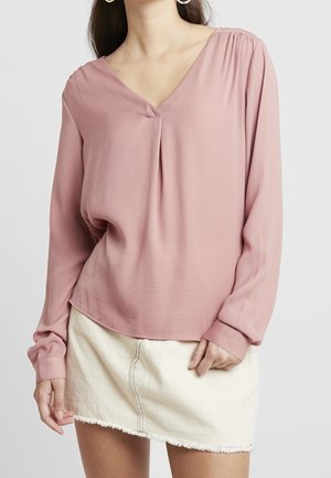 Blusa rosa a maniche lunghe con scollatura a V, spalle arricciate e vestibilità rilassata, abbinata a una gonna di jean mini con orlo sfilacciato.