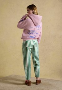 Femme portant un sweat à capuche rose avec un motif de voilier bleu et le texte « Good Vibes », pantalon rayé vert et blanc et chaussures marron, debout de profil.