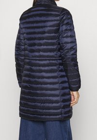 Laivastonsininen toppatakki, jossa vaakasuuntainen tikattu kuvio, korkea kaulus, pitkät hihat ja sileä synteettinen materiaali. Reiden puoliväliin ulottuva, kapean istuvuuden malli.