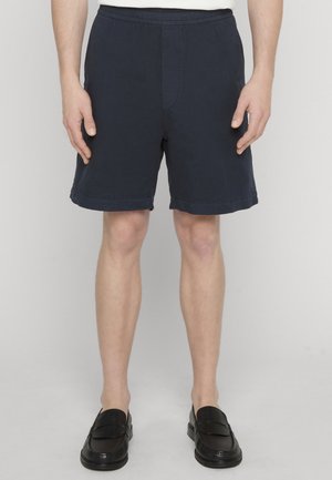Mann iført marineblå shorts, hvit skjorte og svarte loafers, står mot en ensfarget lys bakgrunn.