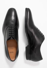 Chaussures habillées en cuir noir présentant une texture lisse, un bout arrondi, un design à lacets et un talon empilé. L'intérieur est beige avec une marque.