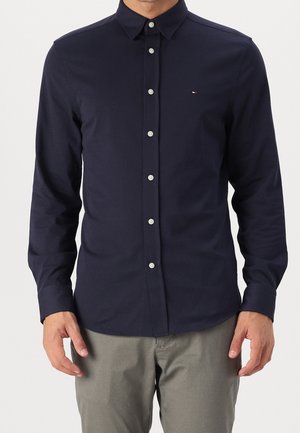 Homme portant une chemise ajustée bleu marine à boutons avec manches longues et un pantalon gris clair, debout devant un fond uni.