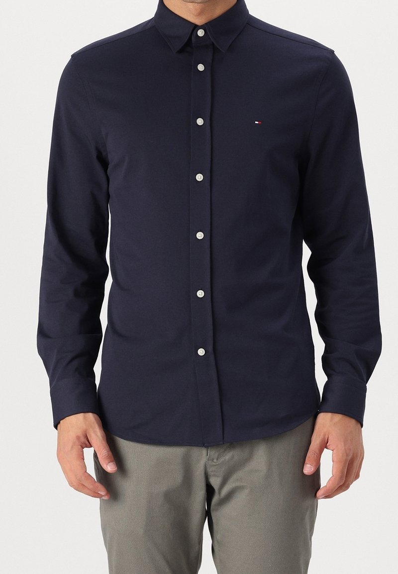 Homme portant une chemise ajustée bleu marine à boutons avec manches longues et un pantalon gris clair, debout devant un fond uni.