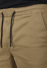 Kaki byxor med svart snörning som har vita diagonala ränder, visar framfickan och detaljerna på midjebandet.