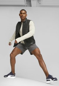 Chaleco acolchado negro sobre una camiseta de manga larga beige, pantalones cortos grises y zapatillas de deporte azul marino. Las texturas suaves y el diseño atlético mejoran la movilidad.