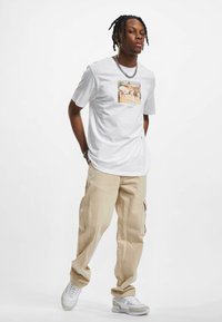 Camiseta de algodón blanca con un estampado gráfico colorido en el centro, combinada con pantalones cargo beige y zapatillas blancas. Colgante de cadena de plata incluido.
