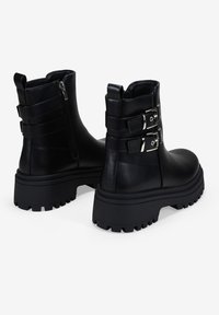Botas de cuero negras de tobillo con suelas gruesas tipo lug, cremalleras laterales y dos correas con hebillas plateadas en cada bota, mostradas desde atrás y de lado.