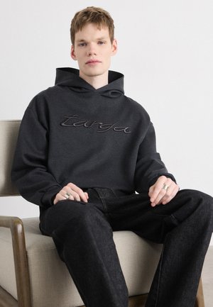 Donkergrijze hoodie met een gestructureerd logo, voorzien van een voorzak in de vorm van een kangoeroe en een capuchon met trekkoord, gecombineerd met donkere spijkerbroeken.