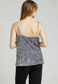 Femme vue de dos portant un haut sans manches en sequins argentés à fines bretelles et un pantalon noir, le tout sur un fond clair.