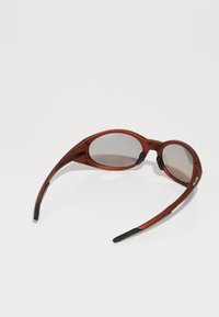 Gafas de sol rojas con montura curva y lentes grises. El diseño presenta un acabado mate, brazos estilizados y acentos de goma negra en las varillas.