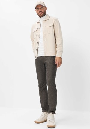 Mand iført beige kasket, beige jakke, hvid rullekrave, mørkegrå bukser og hvide sneakers står mod hvid baggrund.