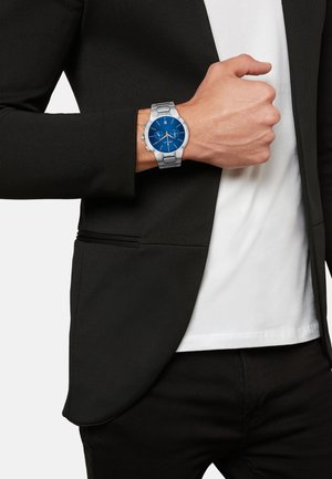 Sølv rustfrit stålur med en blå urskive, der har sølv timeindekser og visere, båret på et håndled, der er delvist synligt i en sort blazer.