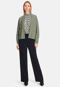 Hellgrüne Strickjacke mit offenem Vorderteil, zwei Fronttaschen, getragen über einem gemusterten Oberteil und kombiniert mit weit geschnittenen schwarzen Hosen und Absatzschuhen.