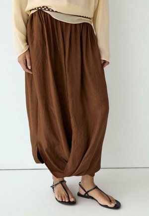 Person trägt lockere braune weite Hose mit gerafftem Saum, beige langärmliges Oberteil und schwarze flache Riemchensandalen auf einem weißen Boden.