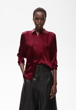 Femme aux cheveux courts portant une chemise en satin bordeaux boutonnée et un pantalon en cuir noir, debout avec une main dans la poche sur fond blanc.
