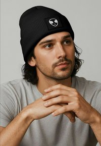 Black ribbed beanie with a square patch featuring an alien graphic. Worn with a light gray t-shirt, model has dark wavy hair.