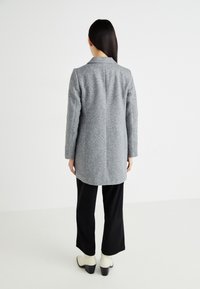 Manteau en mélange de laine gris avec une coupe longue, des revers en pointe et des manches droites, associé à un pantalon noir à jambe large et à des bottines crème à la cheville.