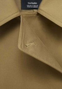 Col roulé en coton beige avec un design pointu, présentant des coutures visibles et un détail de bouton sur le côté près du col.