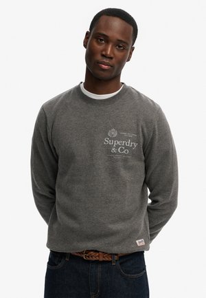 Giovane uomo che indossa una felpa grigia Superdry & Co, una canottiera bianca, una cintura marrone intrecciata e jeans blu scuro, in piedi con le mani dietro la schiena.