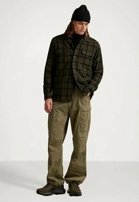 Rutete flanellskjorte i mørkegrønn og svart, kombinert med olivengrønne cargobukser og svarte joggesko. Modellen har på seg en svart lue.