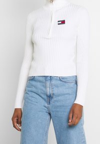 Col roulé blanc côtelé avec une fermeture éclair quart et un petit patch logo. Associé à un jean taille haute bleu clair. Texture douce, coupe ajustée.