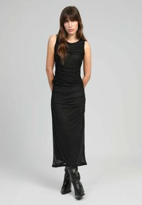 Robe noire sans manches avec fronces, silhouette longue, design ajusté et tissu texturé. Associée à des bottes noires hautes jusqu'aux genoux.