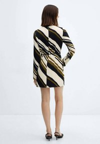 Eine Frau, die von hinten steht, trägt ein langärmliges, knielanges Kleid mit schwarz-weißen und olivgrünen diagonalen Streifen, kombiniert mit schwarzen High Heels.