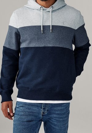 Homme portant un sweat à capuche aux trois tons avec des panneaux horizontaux gris clair, gris moyen et bleu marine, assorti à un jean bleu.