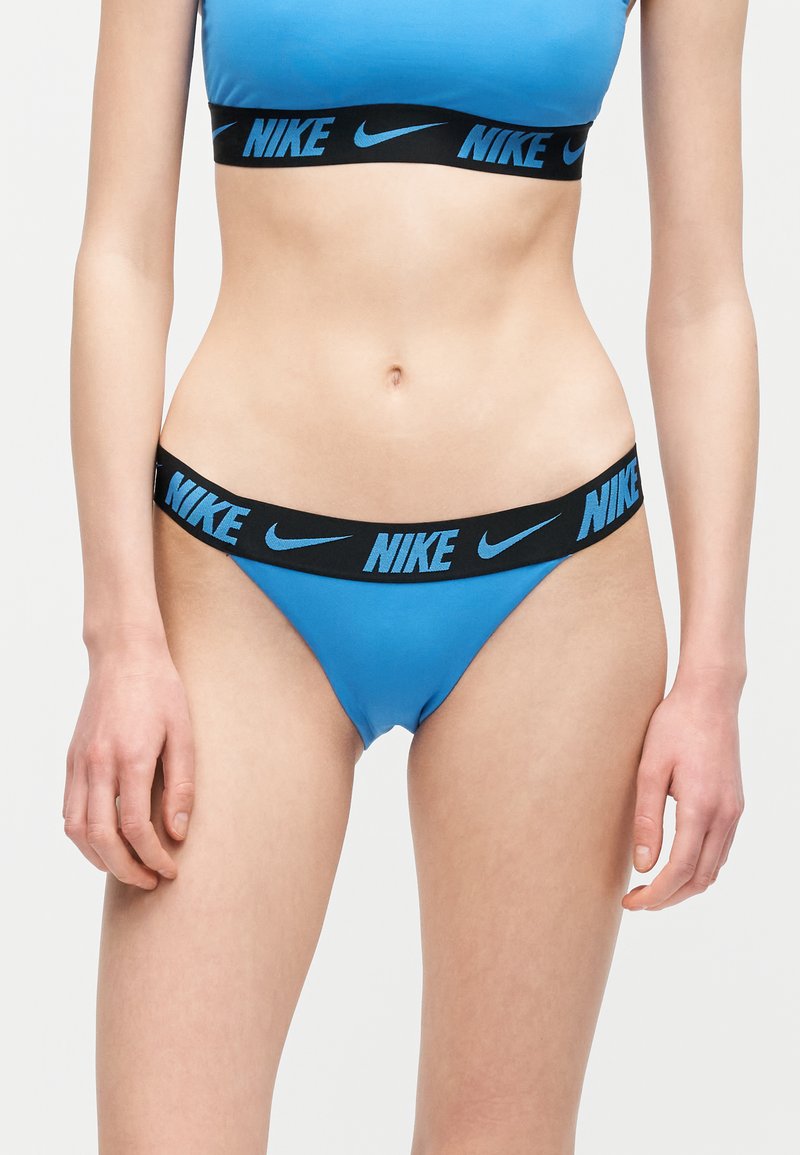 Žena u plavom sportskom grudnjaku marke Nike i odgovarajućem plavom donjem rublju s crnim pojasima na kojima su plavi Nike logotipi.