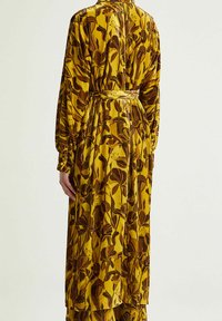 Vestito lungo giallo a fiori con tessuto testurizzato con motivi marroni, vita strette e maniche lunghe. Offre una silhouette fluente ed elegante.