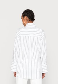 Camicia oversize bianca a righe con colletto a punta, spalle a caduta e maniche lunghe con ampi polsini. La vista posteriore mostra un yoke orizzontale.